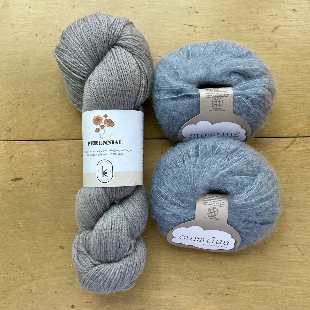 The contents of an "Oslo Hat" Kit - Hillsborough Yarn Shop edition are laid out on a wooden surface: one skein of Kelbourne Woolens Perennial in Fossil and two balls of Fyberspates Cumulus in Mithril