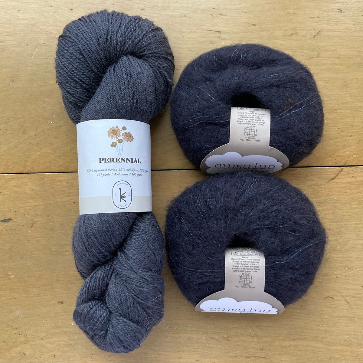 The contents of an "Oslo Hat" Kit - Hillsborough Yarn Shop edition are laid out on a wooden surface: one skein of Kelbourne Woolens Perennial in Lead and two balls of Fyberspates Cumulus in Onyx 