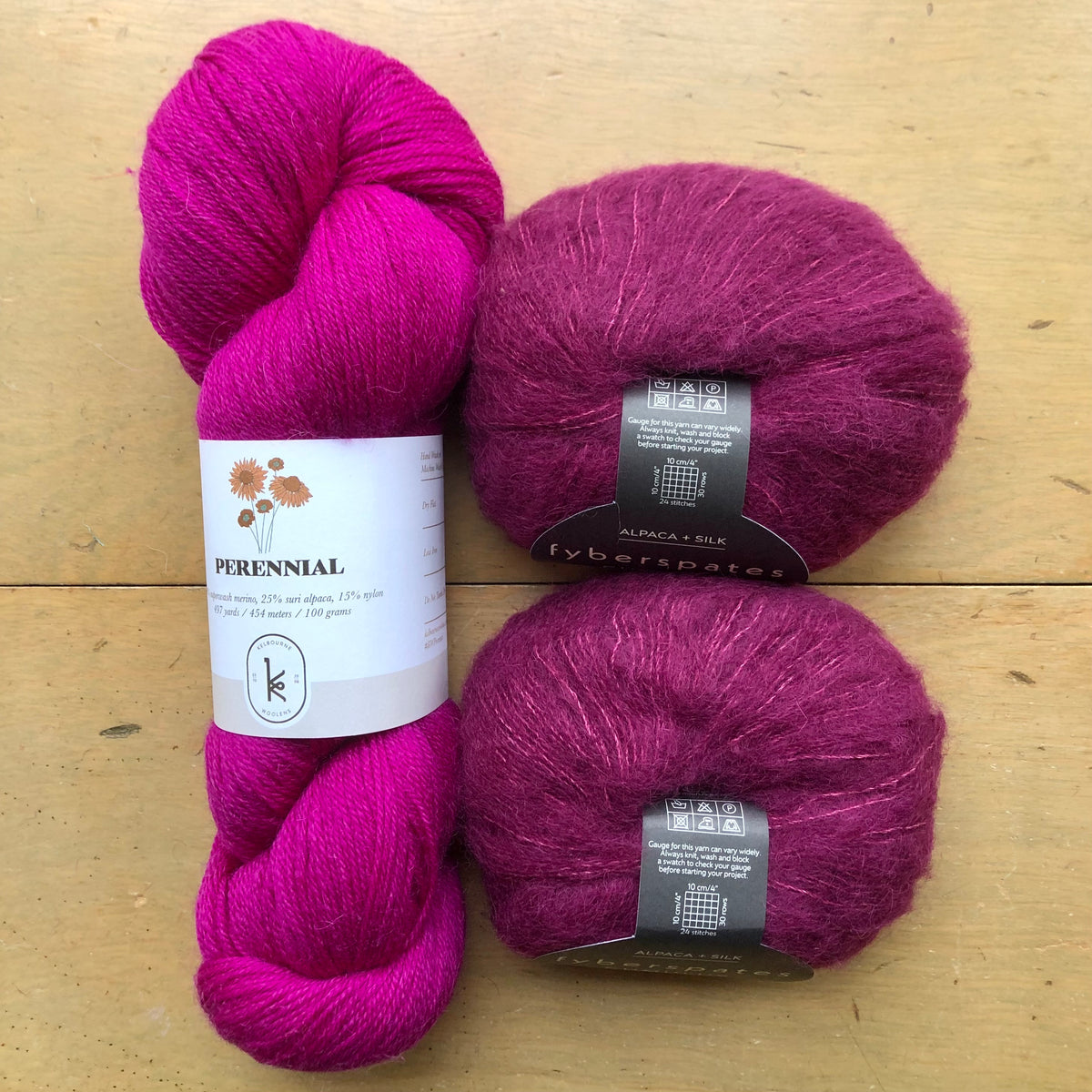 The contents of an "Oslo Hat" Kit - Hillsborough Yarn Shop edition are laid out on a wooden surface: one skein of Kelbourne Woolens Perennial in Raspberry and two balls of Fyberspates Cumulus in Plum. 