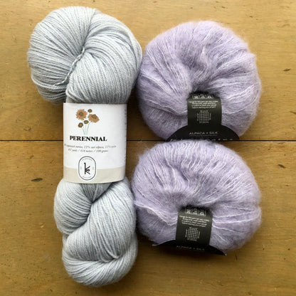 The contents of an "Oslo Hat" Kit - Hillsborough Yarn Shop edition are laid out on a wooden surface: one skein of Kelbourne Woolens Perennial in Silver and two balls of Fyberspates Cumulus in Ethereal. 