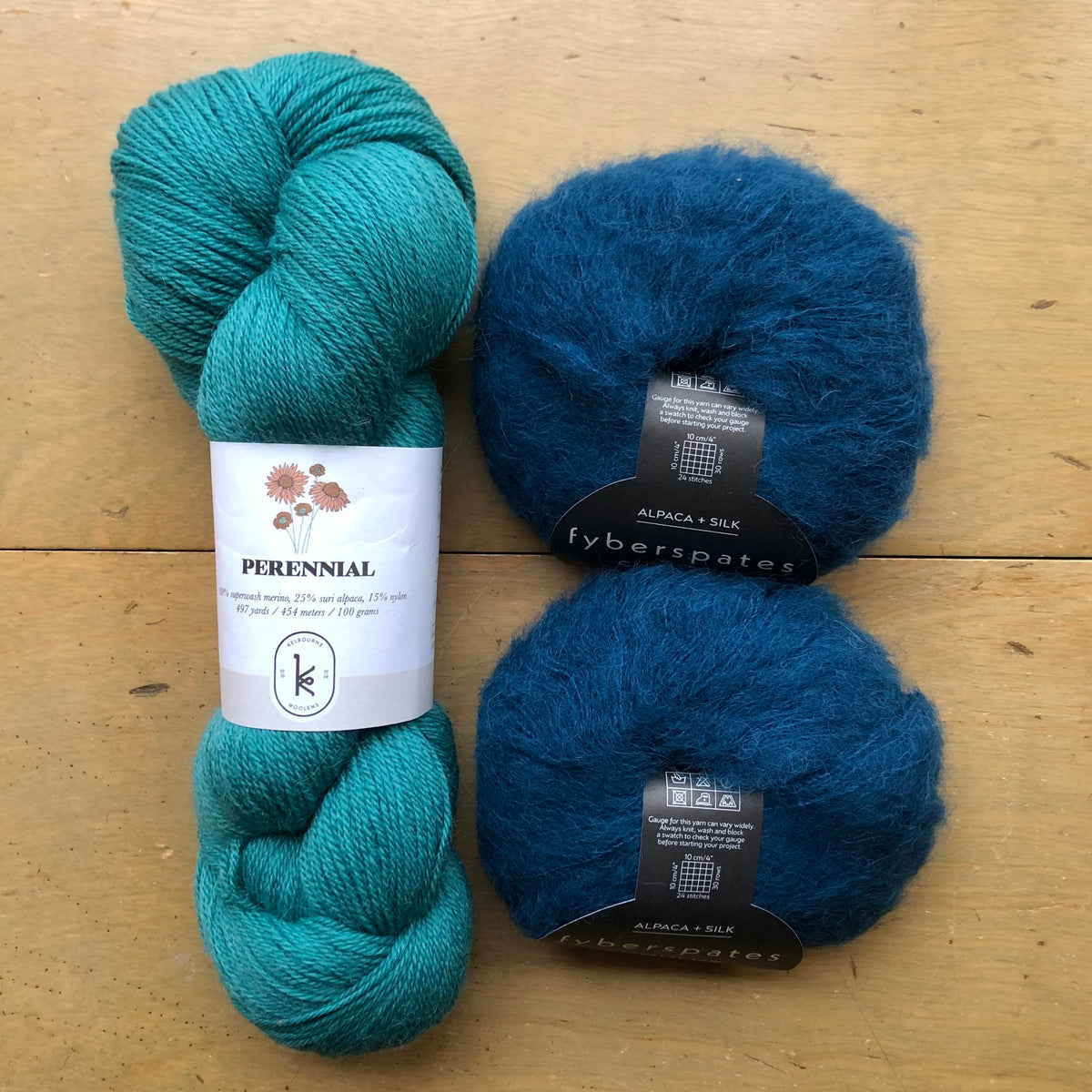 The contents of an "Oslo Hat" Kit - Hillsborough Yarn Shop edition are laid out on a wooden surface: one skein of Kelbourne Woolens Perennial in Teal Green and two balls of Fyberspates Cumulus in Moonlight. 
