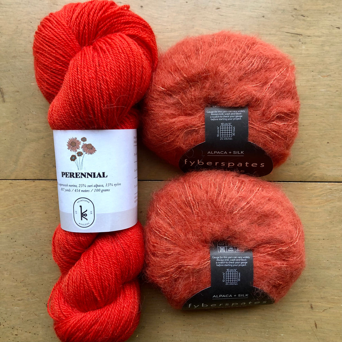 The contents of an "Oslo Hat" Kit - Hillsborough Yarn Shop edition are laid out on a wooden surface: one skein of Kelbourne Woolens Perennial in Vermillion and two balls of Fyberspates Cumulus in Foxy. 