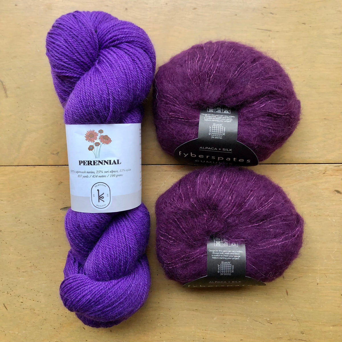 The contents of an "Oslo Hat" Kit - Hillsborough Yarn Shop edition are laid out on a wooden surface: one skein of Kelbourne Woolens Perennial in Violet and two balls of Fyberspates Cumulus in Aubergine. 